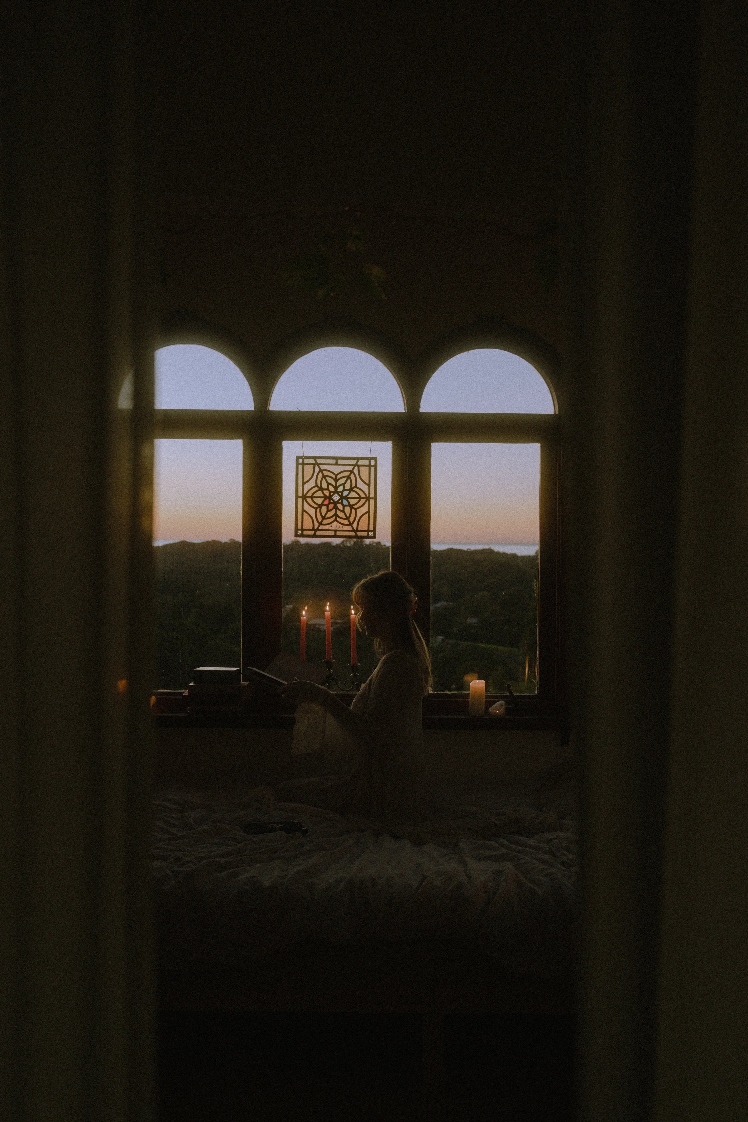 Person sitting by a window with candles, silhouetted against a sunset.